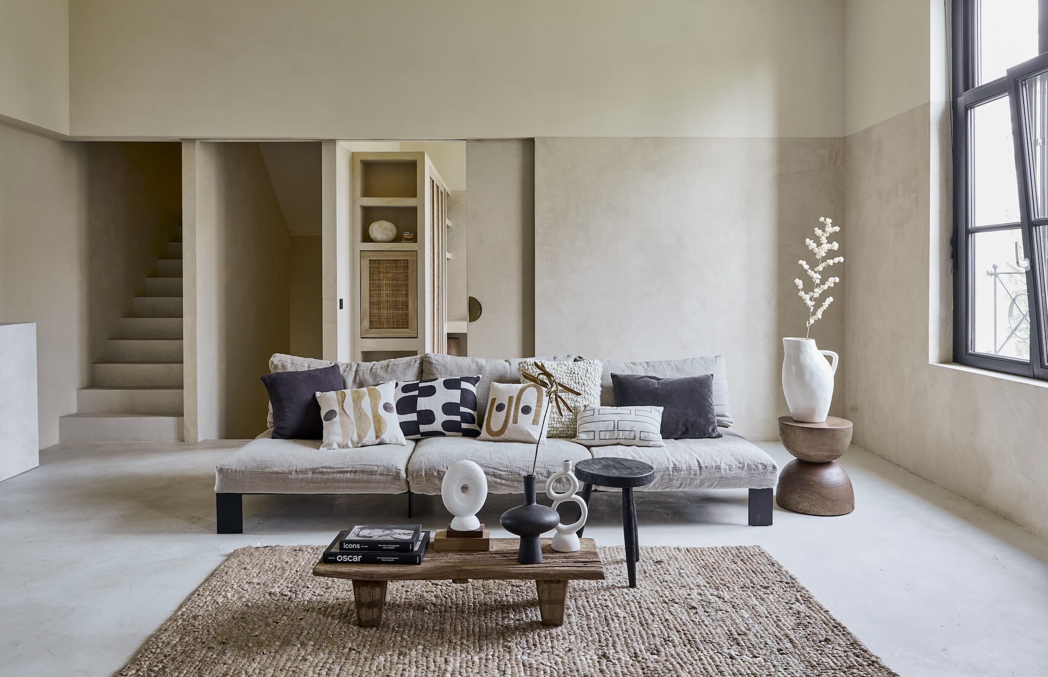 styling interior-styling prop-styling set-design Living room with cement walls and floors couch with light grey pillows, graphic throw pillows brown carpet white vase on wood side table