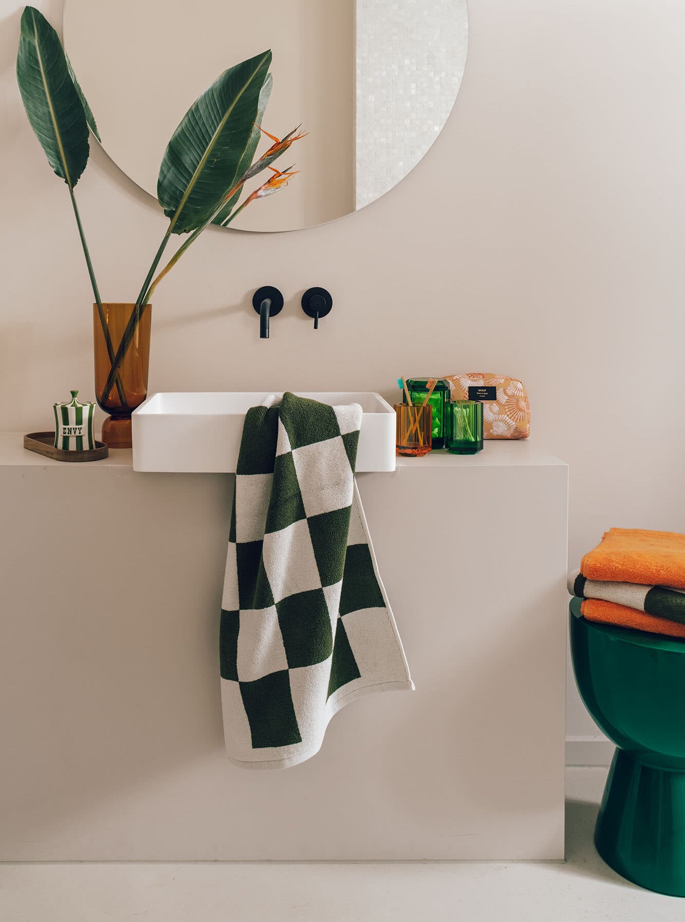 photography still life interior wall mounted sink, black wall faucet, dark green and white checkered towel round mirror green porcelain seat orange towels